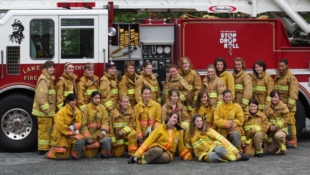 A group of young women standing in front of Fire Truck wearing yellow fire uniforms